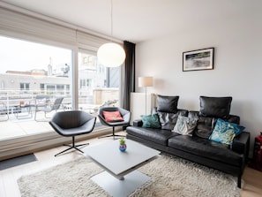 Un salon moderne avec un canapé en cuir noir, une table ronde et une grande fenêtre donnant sur le paysage urbain.
