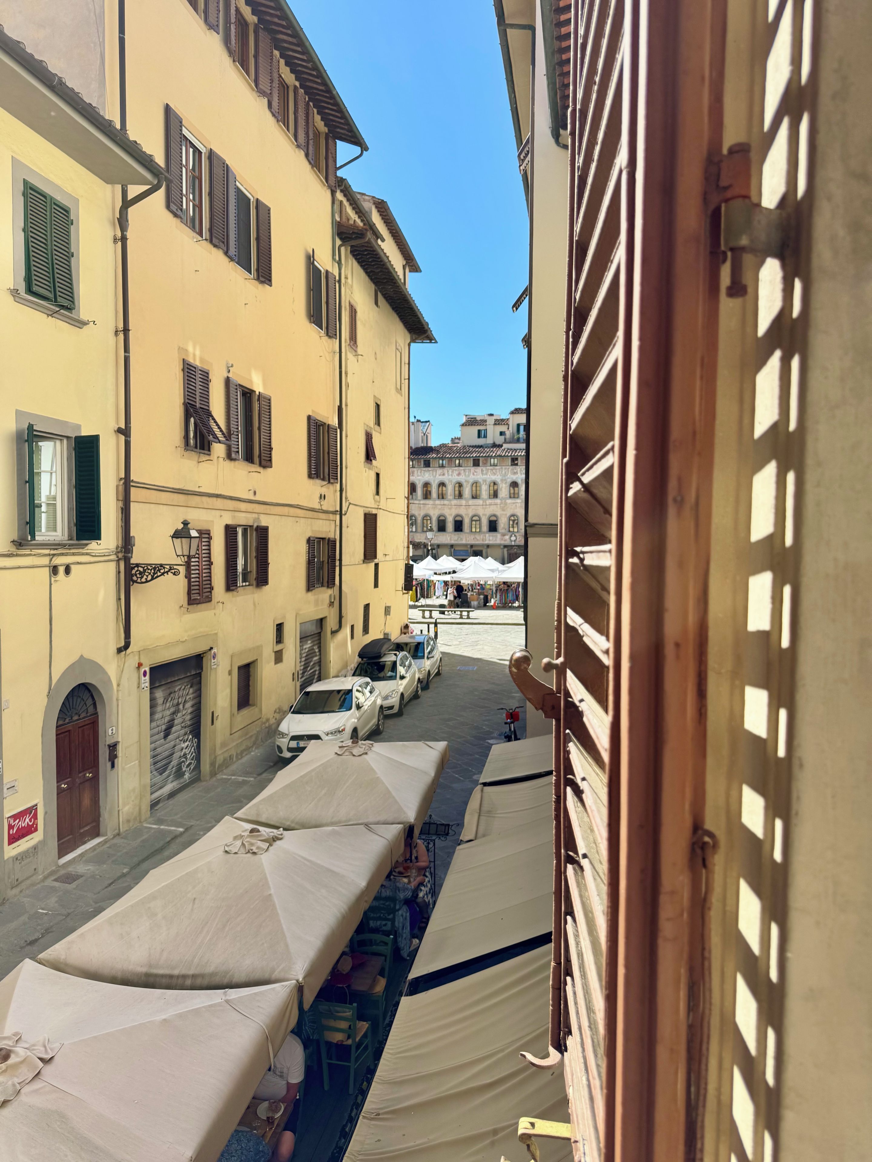 A narrow street with buildings on either side, a row of white tents, and parked cars.