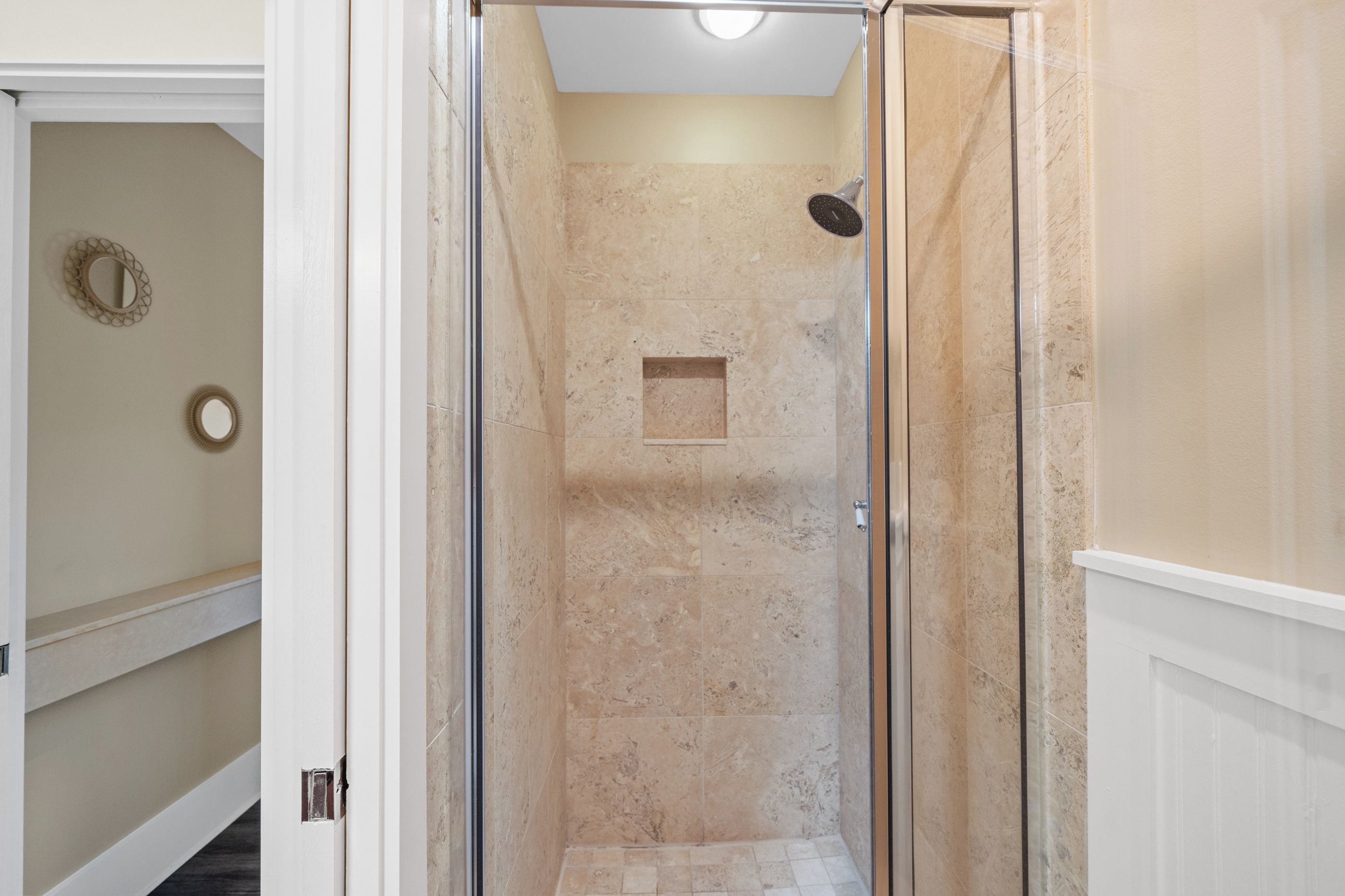 A walk-in shower with a glass door, beige marble walls, and a small square vent.