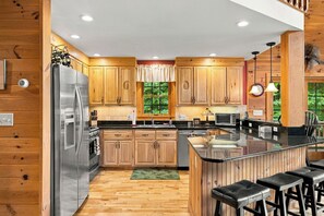 A modern kitchen with wooden cabinets, a stainless steel refrigerator, a built-in oven, and a large island with bar stools. - Luxury Home|Steps from Whiteface|Near Lake Placid (Wilmington)
