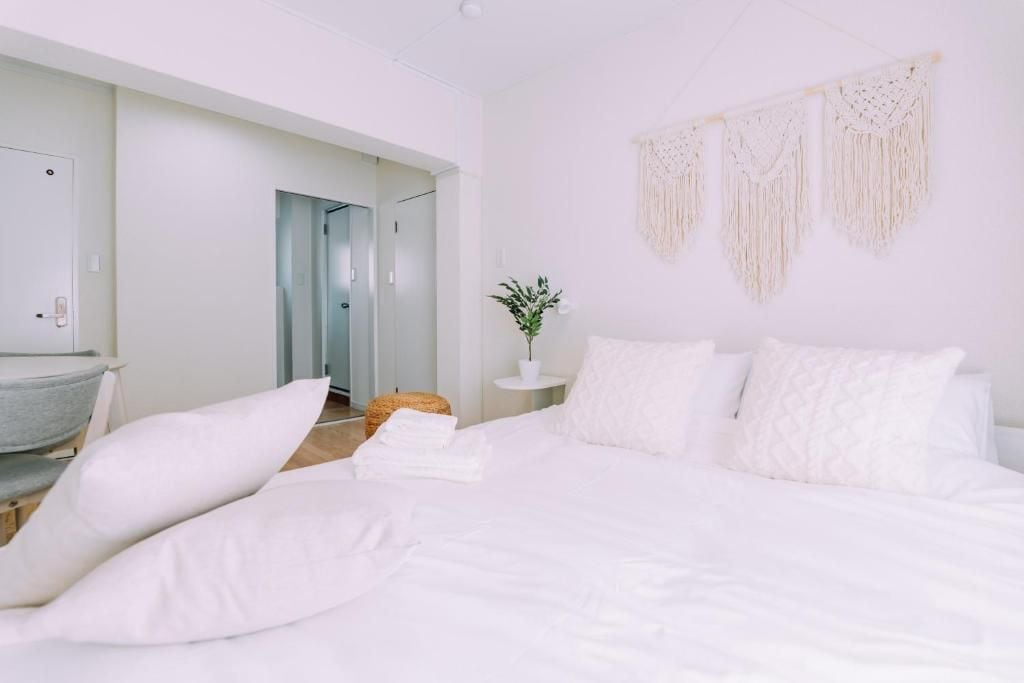 A white-themed bedroom with a bed, pillows, a bedside table, a plant, and a macrame wall hanging.