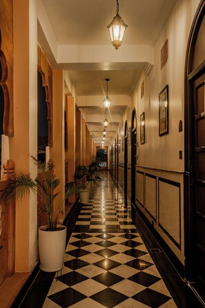 Un corridoio d'hotel con pavimento a scacchi, orologi a muro e una lampada a sospensione.