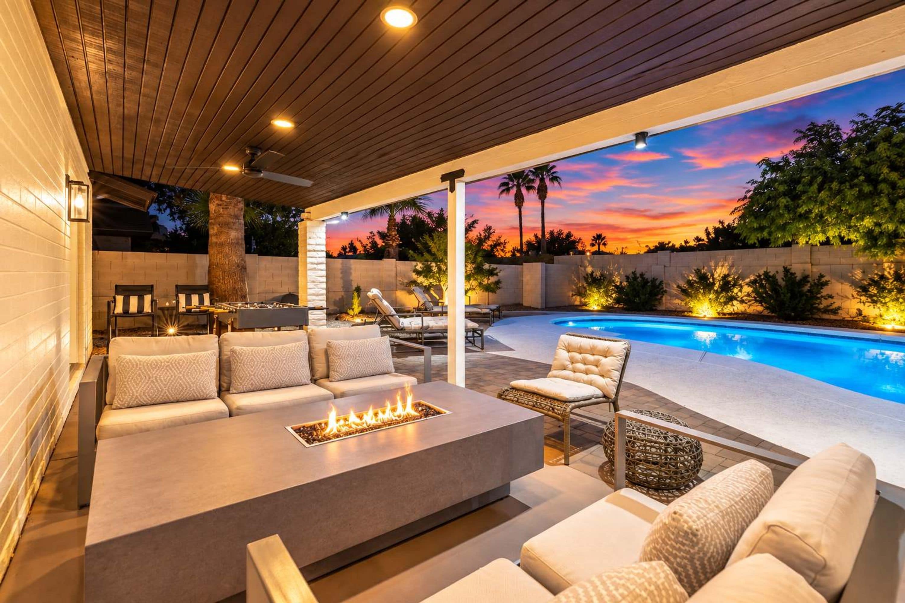 Eine Terrasse im Freien mit Feuerstelle, Sitzgelegenheiten und Blick auf einen Pool bei Sonnenuntergang.
