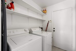 Una sala de bugaderia amb dues rentadores blanques, una estant amb productes de neteja i una porta.