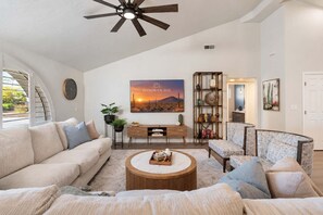 A spacious living room with a high ceiling, a large window, a ceiling fan, a wall-mounted clock, a bookshelf, and a painting of a sunset. - NEW Sonoran Sol Modern Pool Oasis Near Kierland (Scottsdale)