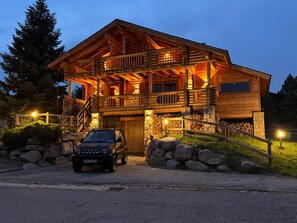 Ein zweistöckiges Blockhaus mit Steinunterbau, ein geparktes Auto davor und eine gut beleuchtete Fassade.