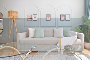 A modern living room with a sofa, a glass coffee table, and a floor lamp. There are framed artworks on the wall and a potted plant. - Luxury1BR in Downtown/Next Dubai Mall & Burj Khalifa (Dubai)