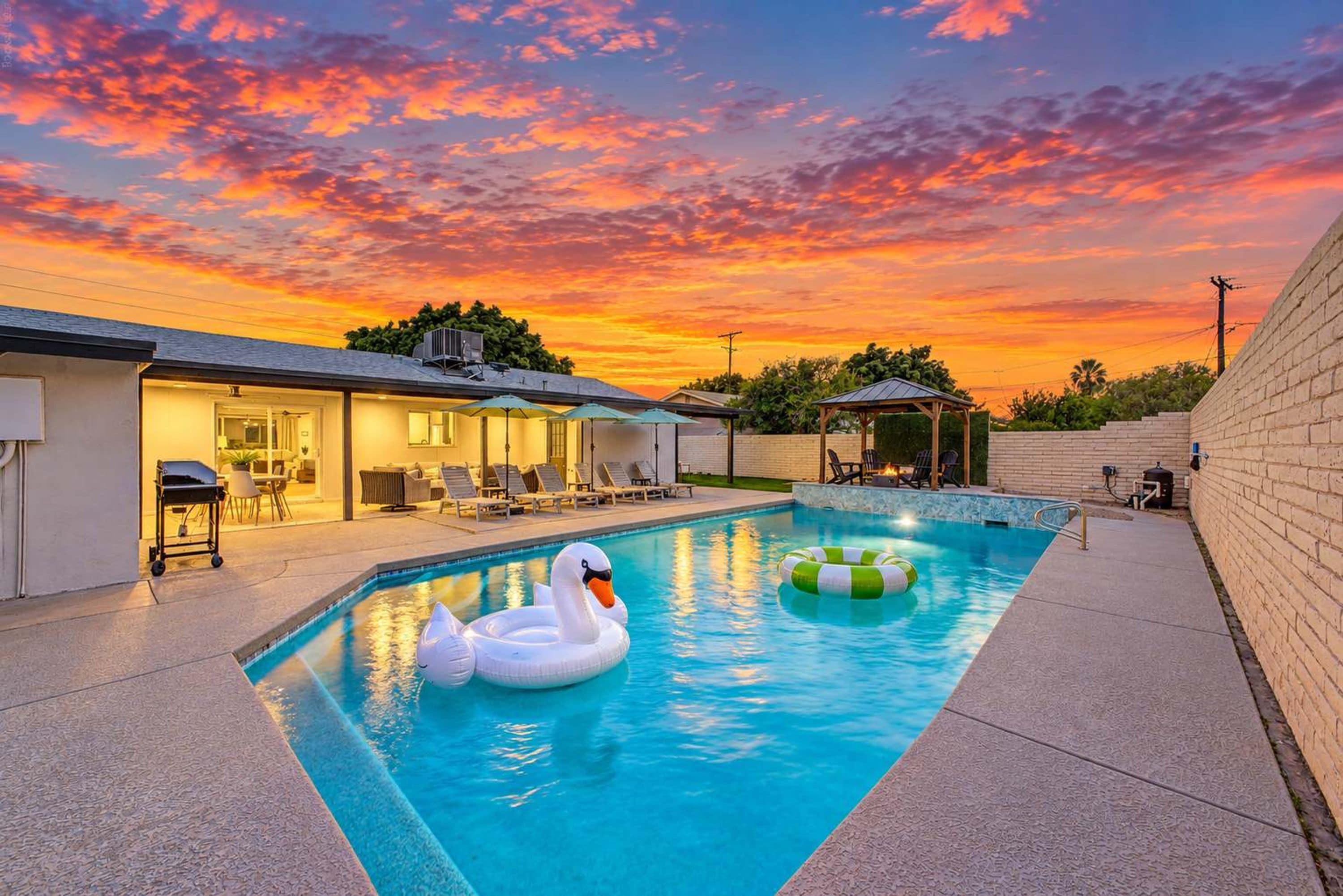 A swimming pool with two swan-shaped floats, a covered seating area, and a sunset view.