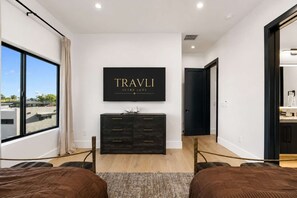 Ein modernes Schlafzimmer mit einem großen Fenster, einer dunklen Holzkommode und einem an der Wand befestigten Fernseher, der 'TRAVLI ULTRA LUXE' zeigt.