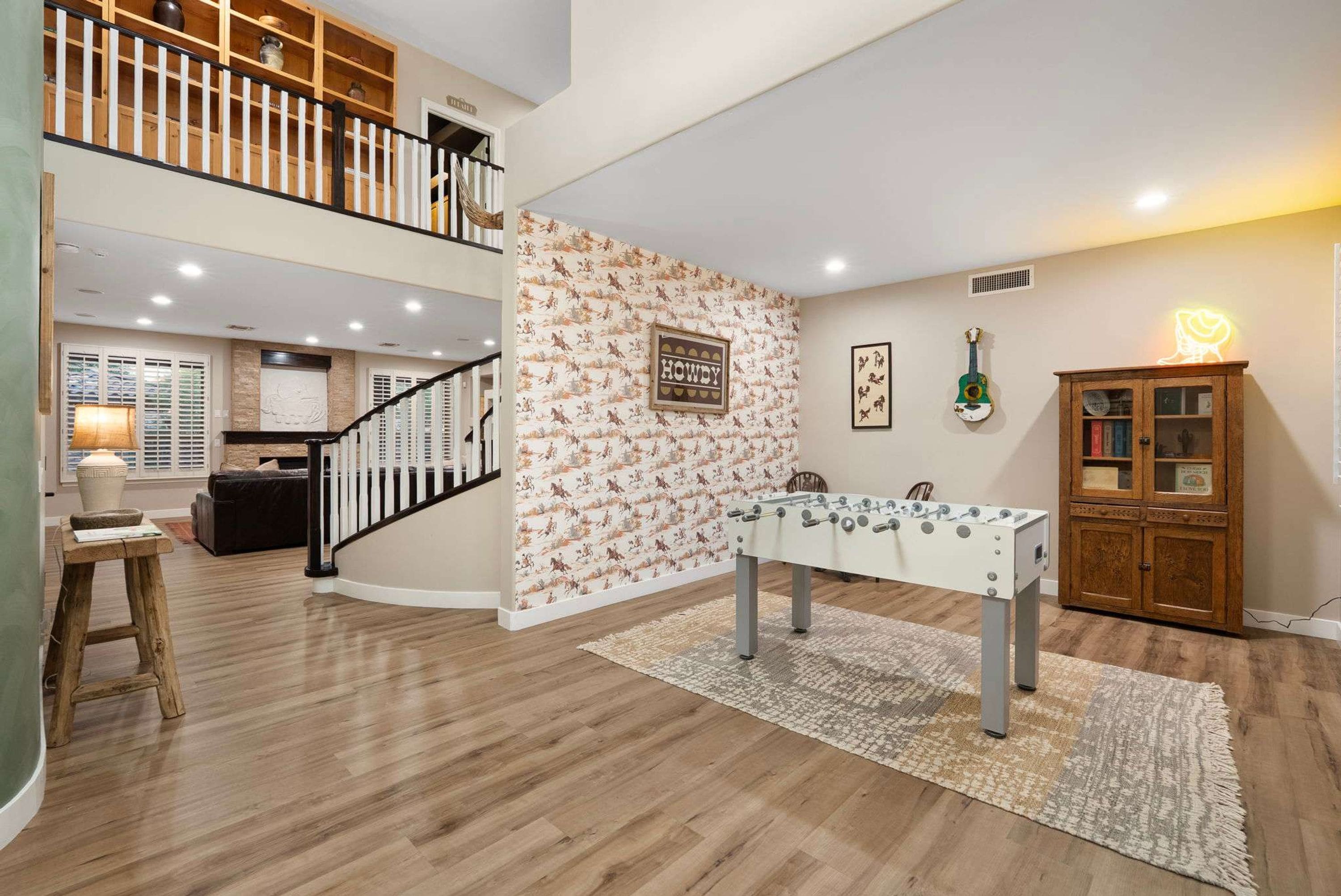 A spacious living area with a high ceiling, a wooden staircase, a foosball table, and a wall with a decorative pattern.