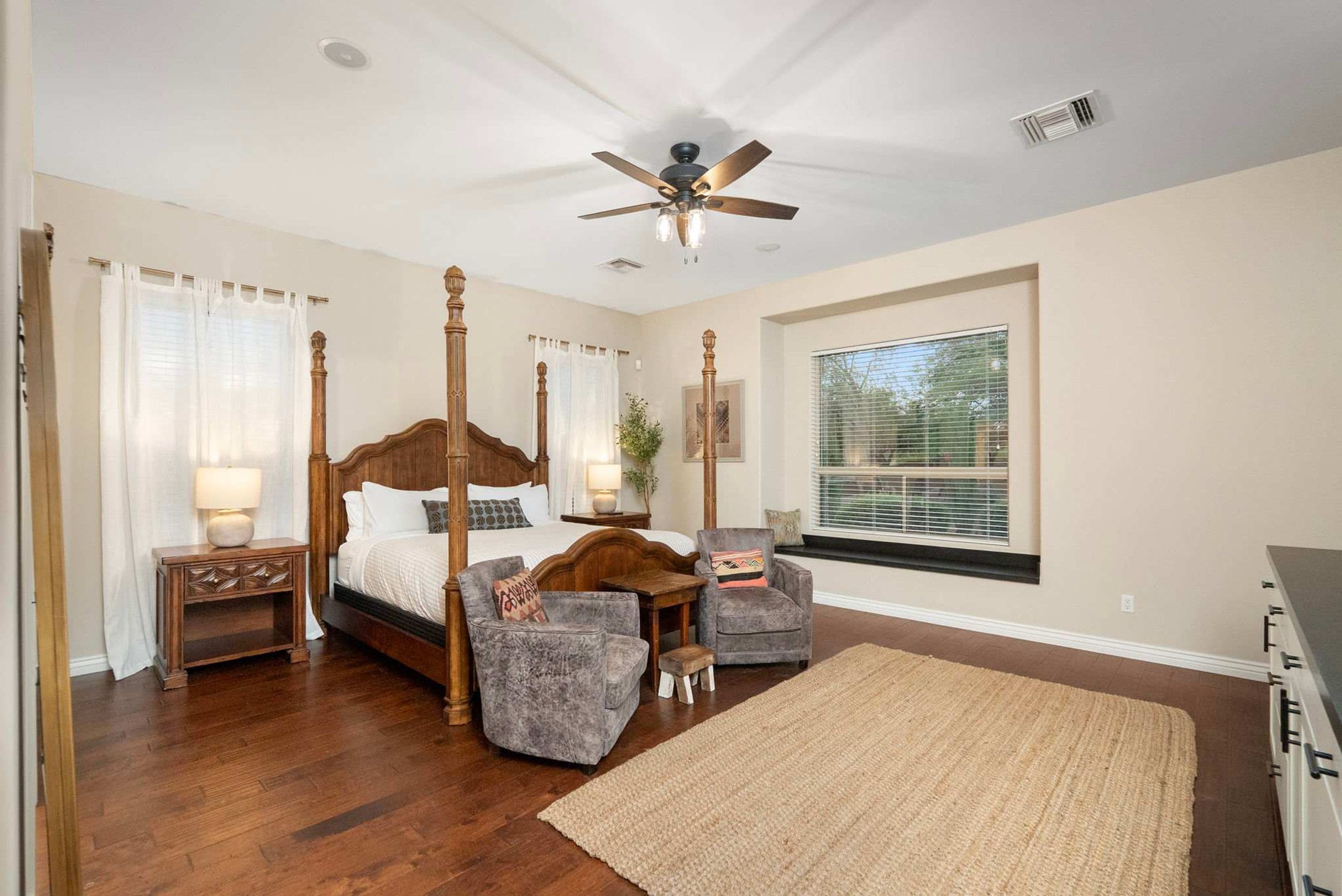 A bedroom with a four-poster bed, two armchairs, a large window with blinds, a ceiling fan, and a wooden floor.