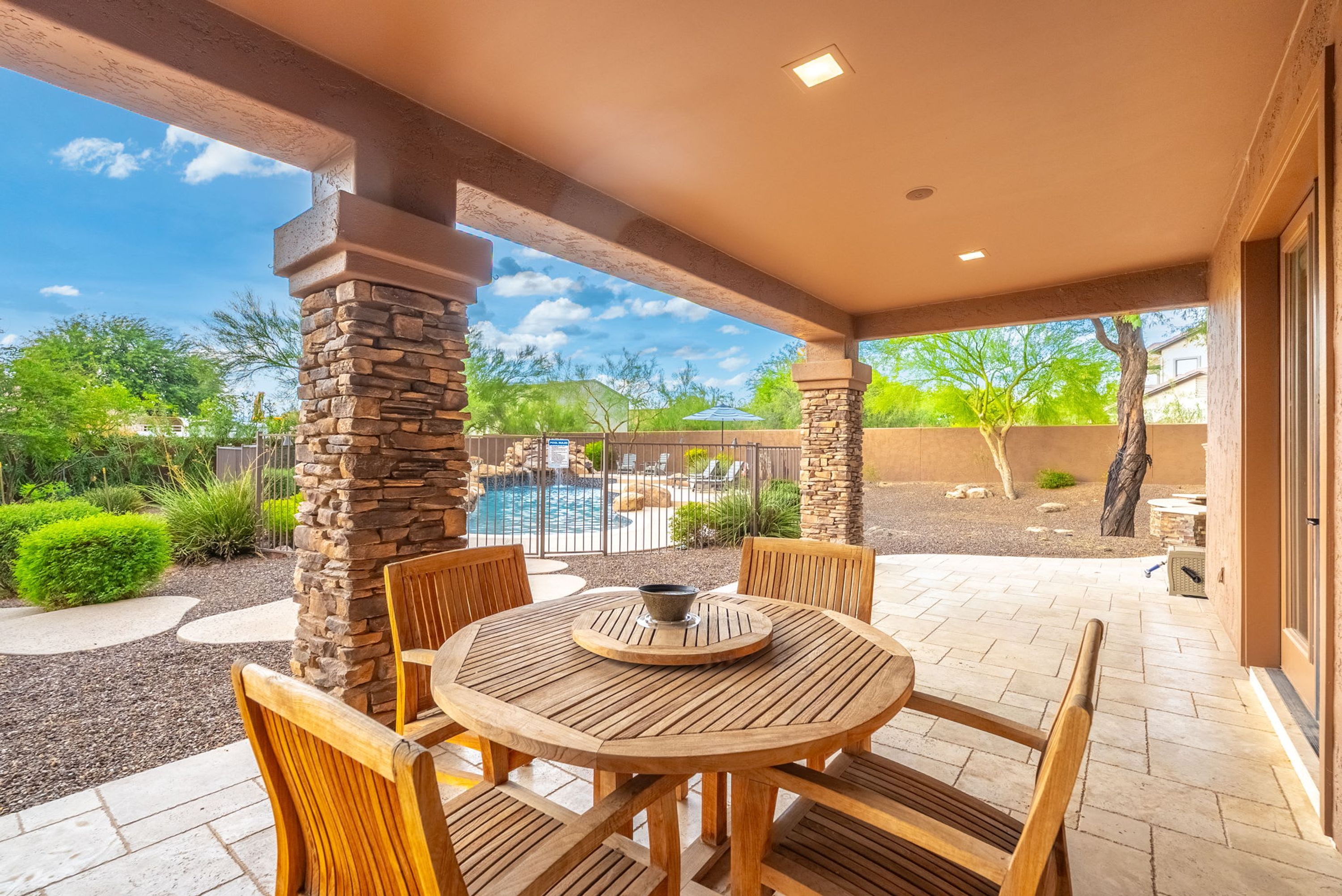 An outdoor patio with a round wooden table and chairs, stone pillars, and a view of a swimming pool.
