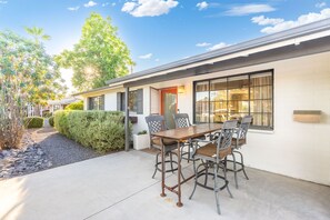 A residential building with a patio area featuring a table and chairs, a window with curtains, and a hedge. - NEW Desert Bloom Pool Spa Game Room (Scottsdale)