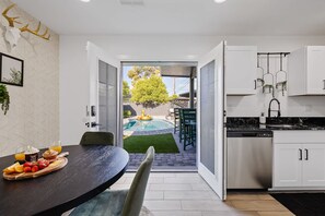 Une cuisine moderne avec des meubles blancs, un coin repas doté d’une table sombre, et une vue sur un jardin avec une piscine et un arbre.