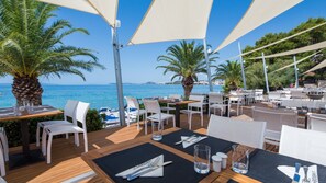 Een buitenterras met tafels en stoelen, palmbomen en uitzicht op zee.