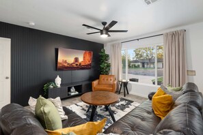 A modern living room with a dark wall, a large painting, a ceiling fan, a sofa with cushions, and a view through a large window. - NEW The Tipsy Cactus Old Town Oasis Pool Bar Putt (Scottsdale)