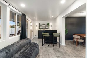 A modern living space with a grey sofa, a dining area with two chairs, a kitchen counter, and a large window with curtains. - NEW The Tipsy Cactus Old Town Oasis Pool Bar Putt (Scottsdale)