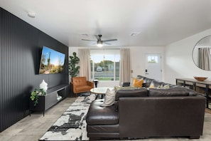 Een moderne woonkamer met een hoekbank, een salontafel, een aan de muur bevestigde televisie en een plafondventilator.