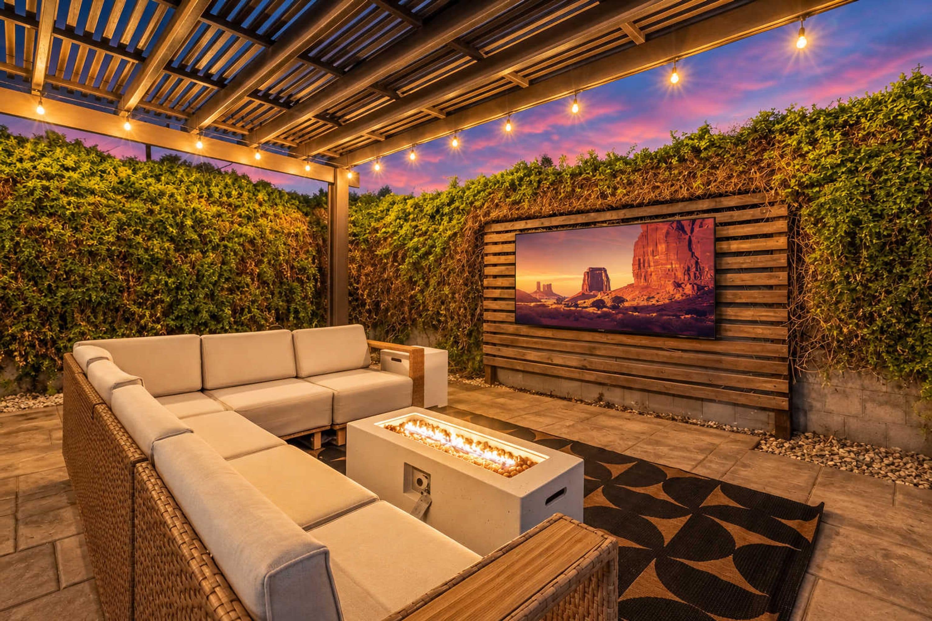 An outdoor seating area with a sofa and a fire pit, surrounded by a wooden pergola and a wall-mounted TV.