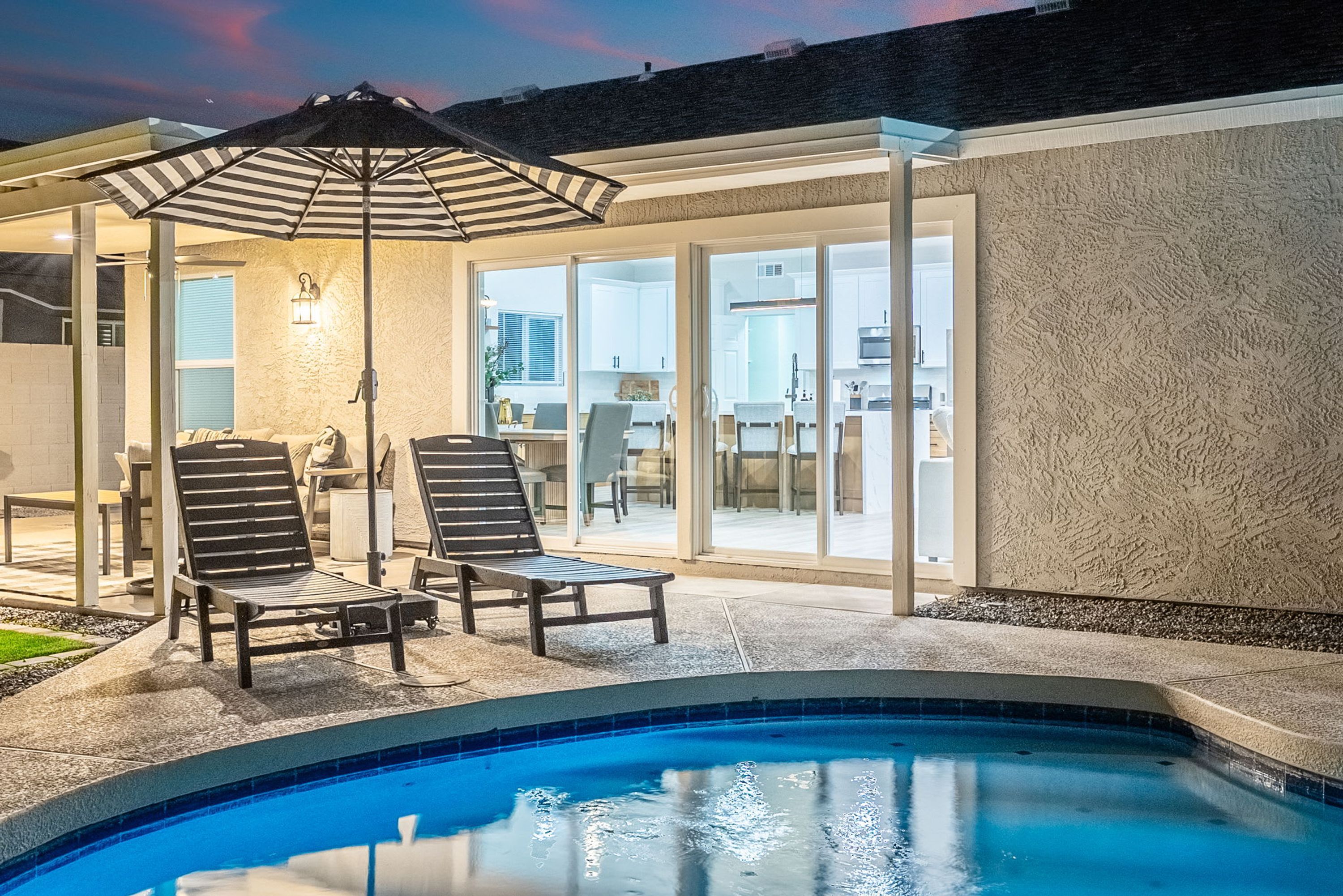 Une piscine dotée de chaises longues et d’un parasol, entourée d’une terrasse d’où l’on peut admirer une cuisine et une salle à manger.