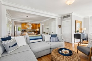 Ein modernes Wohnzimmer mit einem grauen Ecksofa, einem runden Couchtisch und einem Essbereich mit Holzschränken sowie einer Küche im Hintergrund.