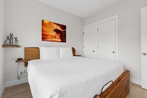 Ein Schlafzimmer mit einem großen Bett, einem Holzkopfbrett, einem Nachttisch mit Telefon, einem Wandregal mit einer Skulptur und einer Tür mit Griff.