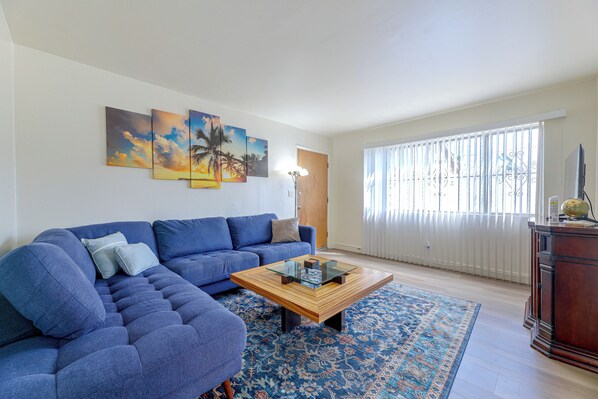 A living room with a sectional sofa, a coffee table, a patterned rug, and a large window with blinds. - Quaint & Cozy Hideaway! 15 Min to LAX (Los Angeles)