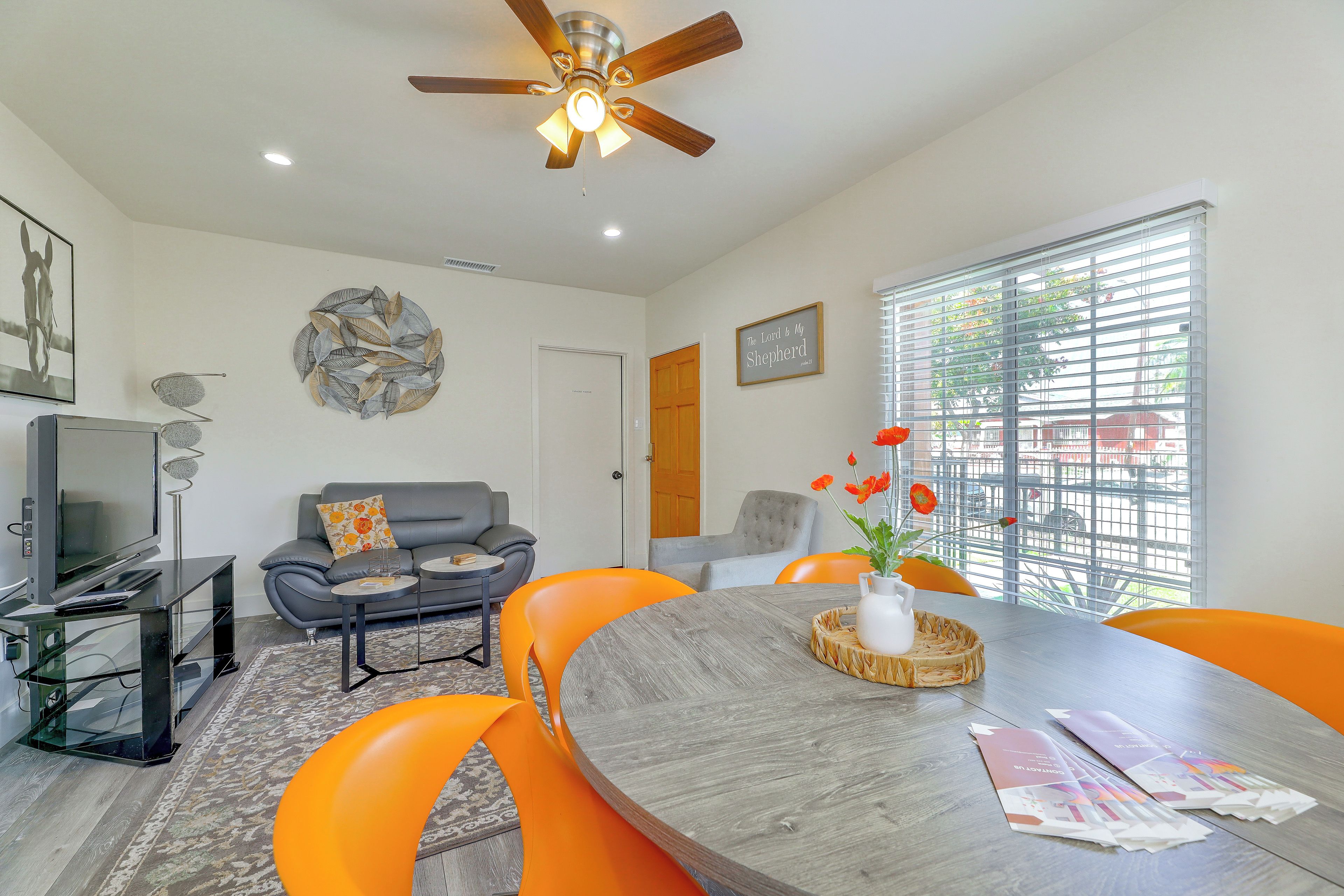 A modern living room with a ceiling fan, a flat-screen TV, a grey sofa, orange chairs, a dining table, and a window with blinds.