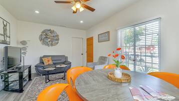 A modern living room with a ceiling fan, a flat-screen TV, a grey sofa, orange chairs, a dining table, and a window with blinds.