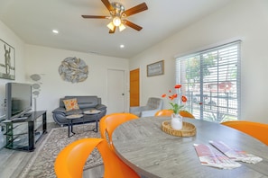 A modern living room with a ceiling fan, a flat-screen TV, a grey sofa, orange chairs, a dining table, and a window with blinds. - LA Home w/ Porch, 3 Mi to Stadiums! (Los Angeles)