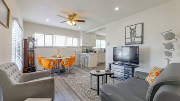 A modern living room with a sofa, a coffee table, and a television. There is a dining area with orange chairs and a table. The room has a ceiling fan and a large window with blinds.