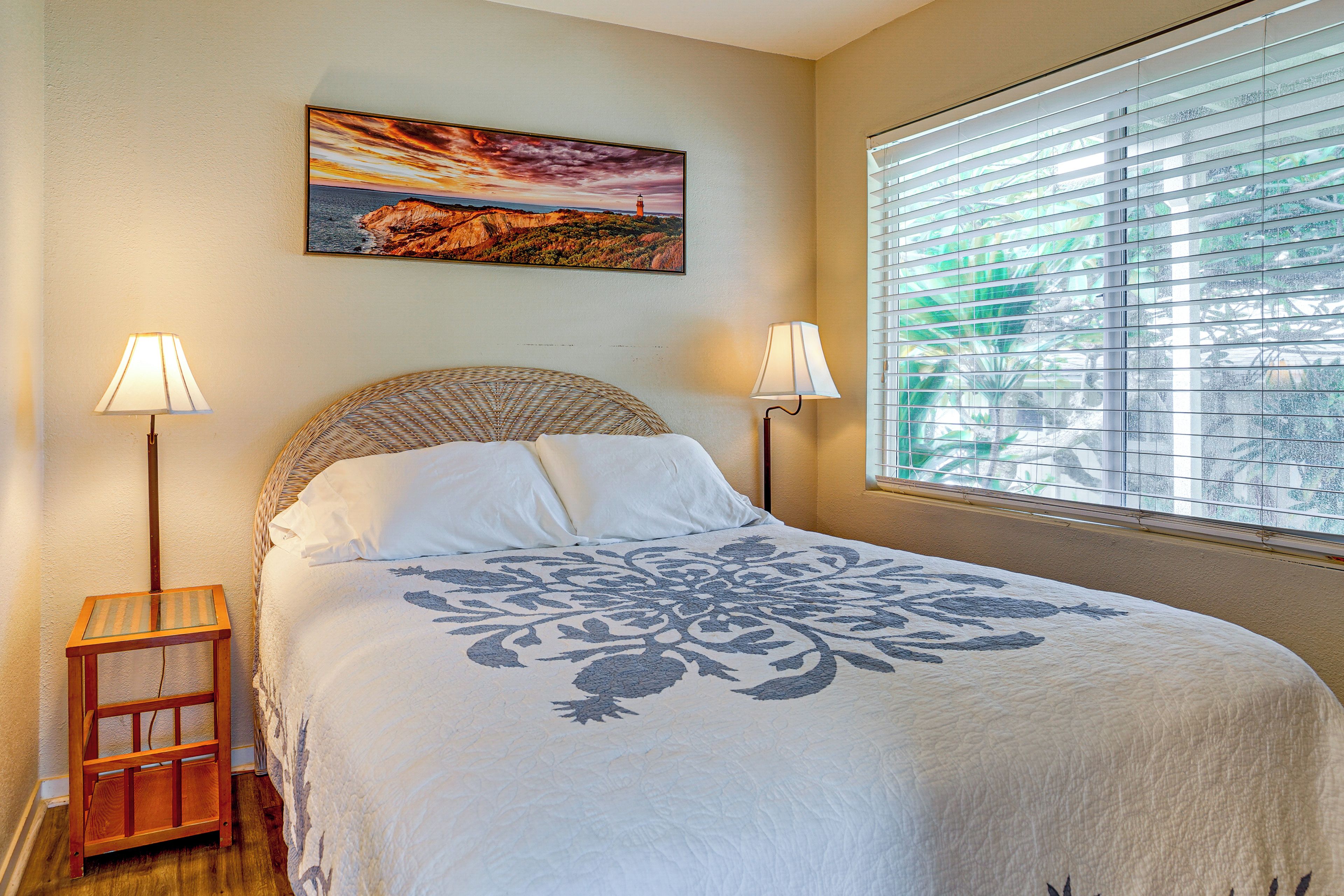 A bedroom with a bed, two bedside lamps, a window with blinds, and a painting on the wall.