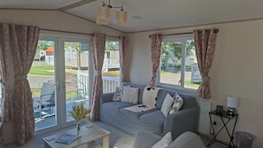 A living room with a grey sofa, a small coffee table, and a vase with flowers. There are curtains on the windows, and a view of the outside.
