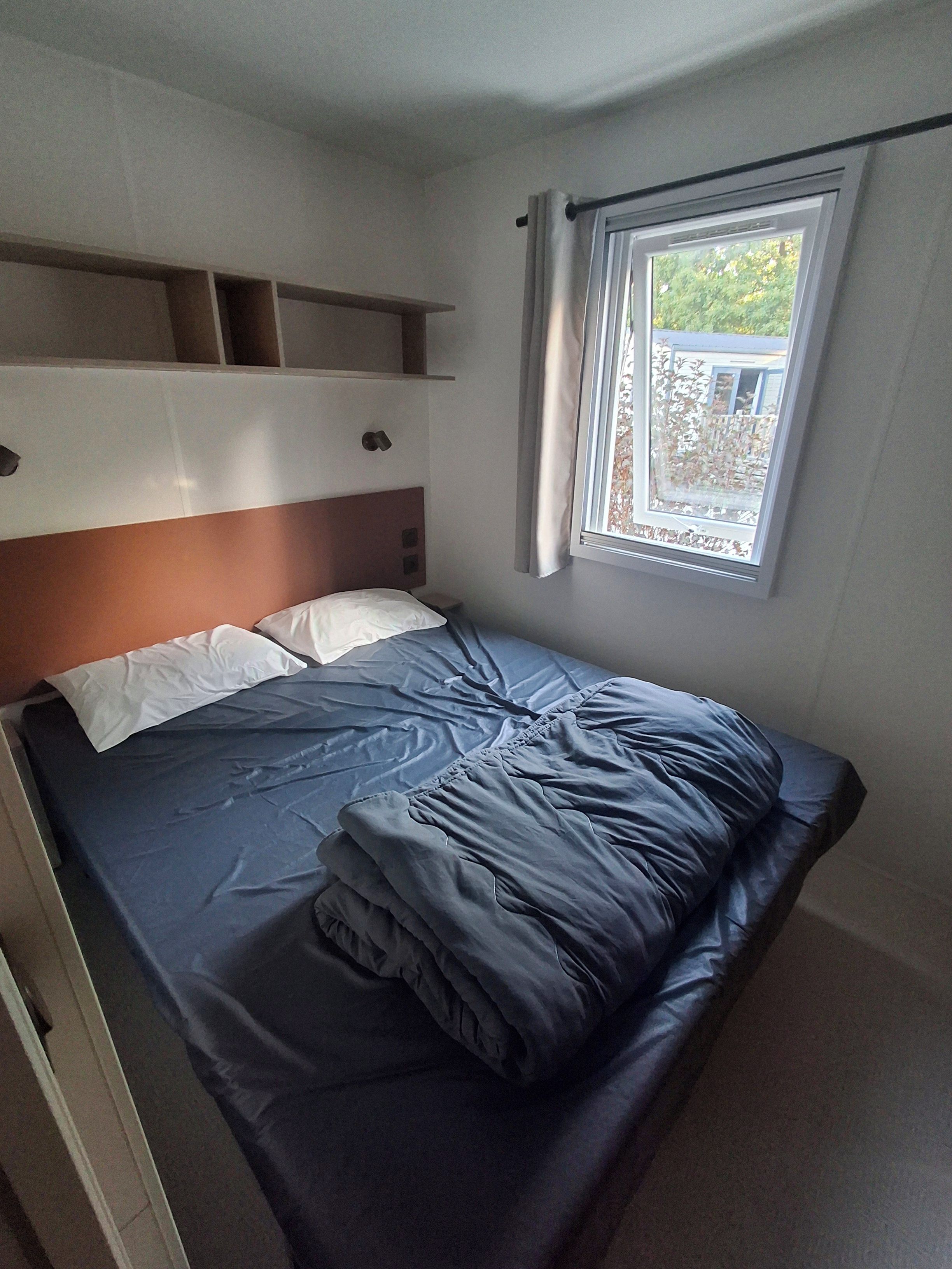 Una cama individual con cabecera, una ventana con cortinas y una repisa sobre la cama.