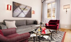 Un salon avec un canapé gris, un fauteuil rouge, une table basse en verre ornée d’un vase et d’une bouteille de vin, et un grand tableau abstrait accroché au mur.