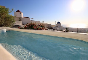 A swimming pool with clear blue water, surrounded by white buildings with blue domes, and a view of the sea in the background. - Ambrosia Cave Villa with Seaview & Private Pool (Exo Gonia)