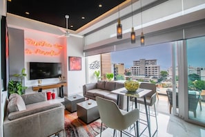 A modern living room with a flat-screen TV, a sofa, a coffee table, and a dining area with chairs and a table. - Romantic Zone Penthouse with Partial Ocean Views (Puerto Vallarta)
