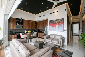 A modern living room with a sofa set, a coffee table, and a dining area with chairs and a kitchen in the background. - Romantic Zone Penthouse with Partial Ocean Views (Puerto Vallarta)