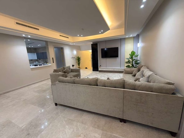 A modern living room with a sectional sofa, a flat-screen TV mounted on the wall, and a kitchen visible through an open doorway. - Aurion Executive Apartment- Rashid Mall (Madinah)