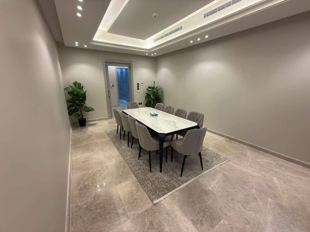 A conference room with a rectangular table, multiple chairs, a patterned rug, and two potted plants.