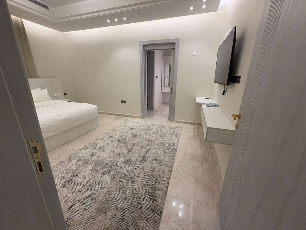 Un dormitorio con una cama, una mesita de noche, un televisor montado en la pared y una puerta que da a otra habitación.