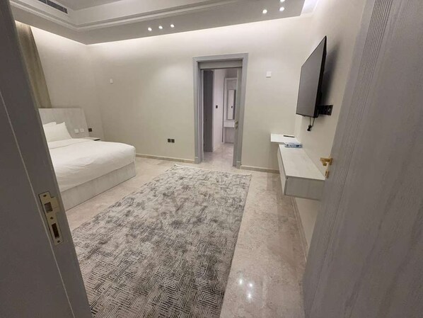 A bedroom with a bed, a bedside table, a TV mounted on the wall, and a door leading to another room.