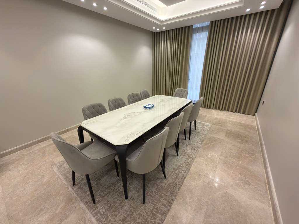 Un salón de conferencias con una mesa de mármol y seis sillas, un ventanal con cortinas y una lámpara empotrada en el techo.