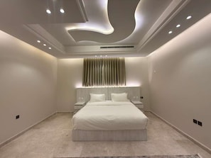 A modern bedroom with a large bed, recessed ceiling lighting, and a decorative wall panel. - Aurion Executive Apartment- Rashid Mall (Madinah)