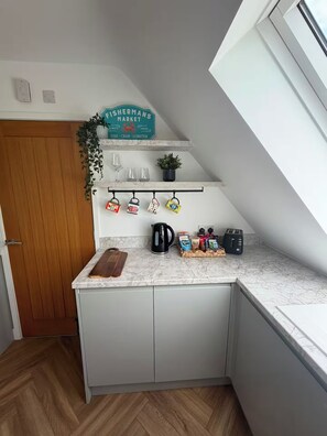 A kitchen with a white countertop, a coffee maker, a kettle, and a toaster. There is a wooden door and a plant on the wall. - ‘The Loft’ Near Whitstable, Parking, EV, Courtyard (Yorkletts)