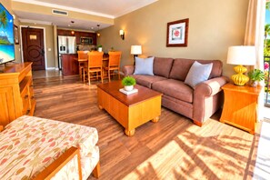 A hotel room with a sofa, a wooden coffee table with a plant, a dining area with chairs, a kitchen, and a large window with curtains. - Honua Kai | Mountain View 1 BR | Free Car w/ 6+ Nights | HKK-230 (Lahaina)
