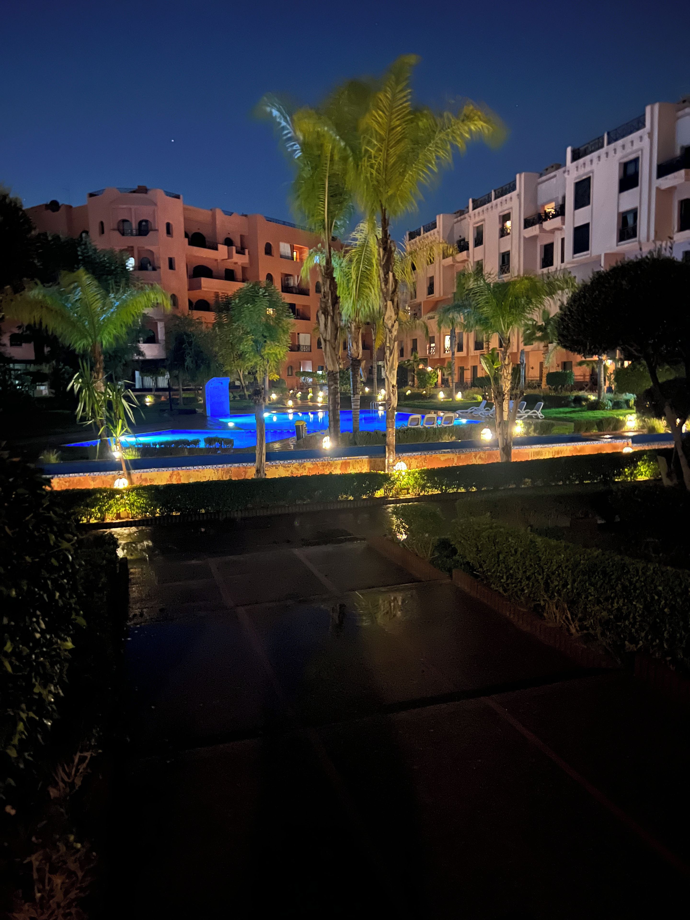 A nighttime view of a hotel with a swimming pool, palm trees, and illuminated pathways.