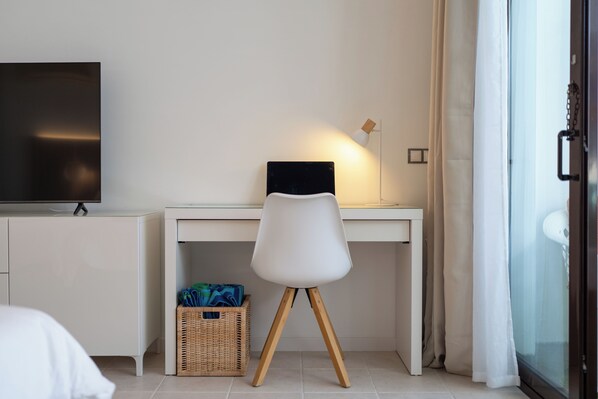 A modern hotel room with a white desk, a flat-screen TV, a white chair, a wicker basket, and a glass door with curtains. - Brand-new duplex with sea views. (CN)