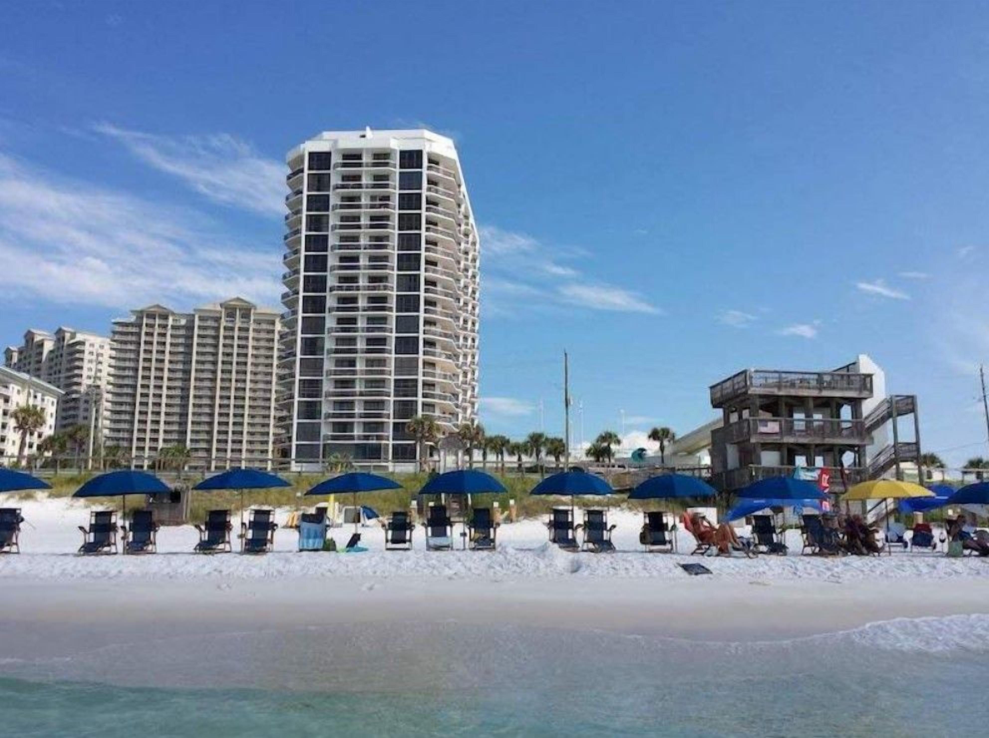 Un hôtel situé en bord de mer, avec plusieurs étages, des parasols et des chaises longues sur la plage.