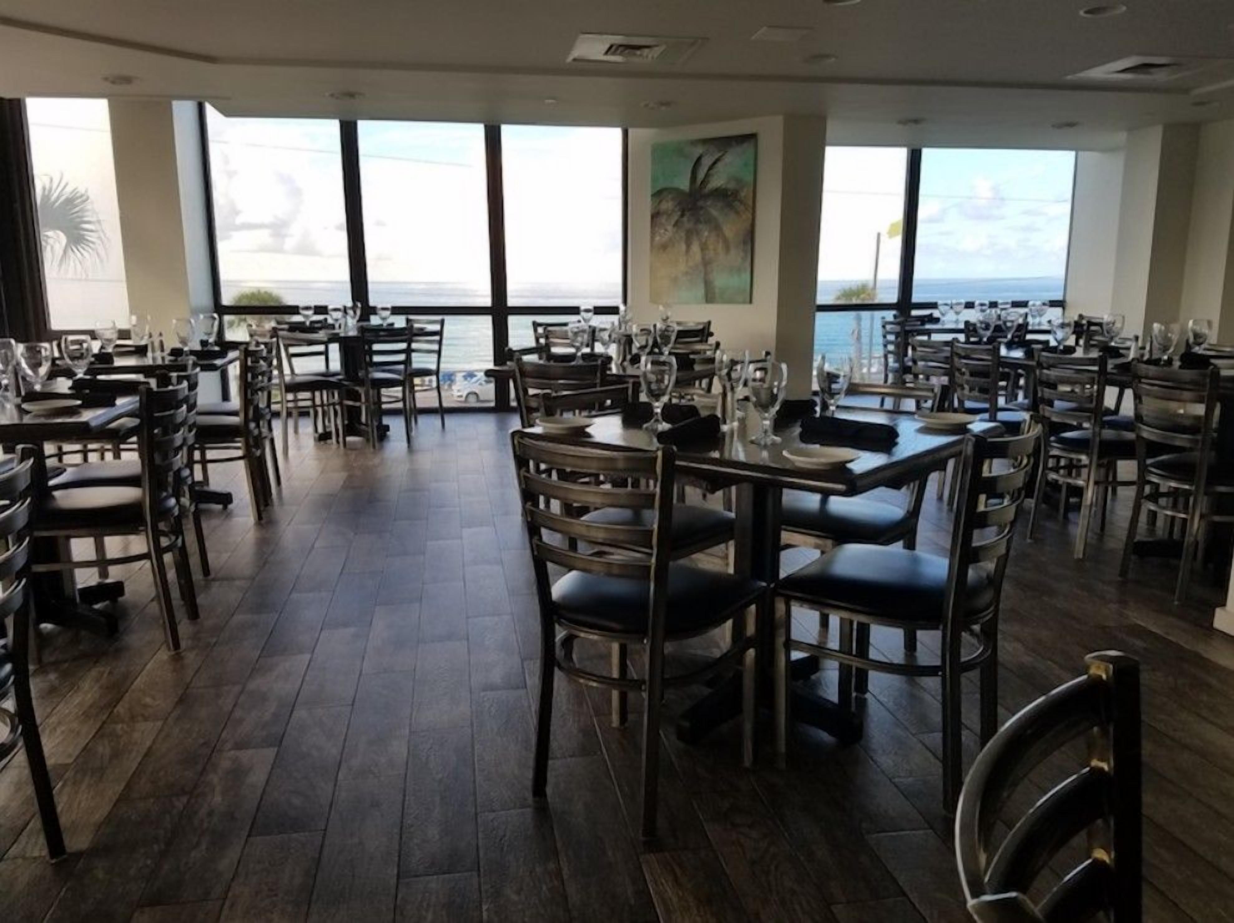 A restaurant with multiple tables and chairs, large windows with a view of the sea, and a modern interior design.